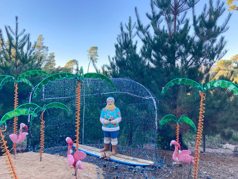 A festive outdoor Christmas display featuring a beach-themed Santa on a surfboard, pink flamingos, palm tree lights, and twinkling decorations among pine trees.