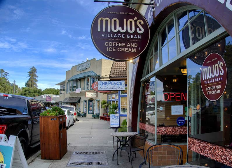 A cozy street scene in Cambria, California, with the exterior of Mojo’s Village Bean coffee shop, its inviting outdoor seating, and a row of boutique shops.