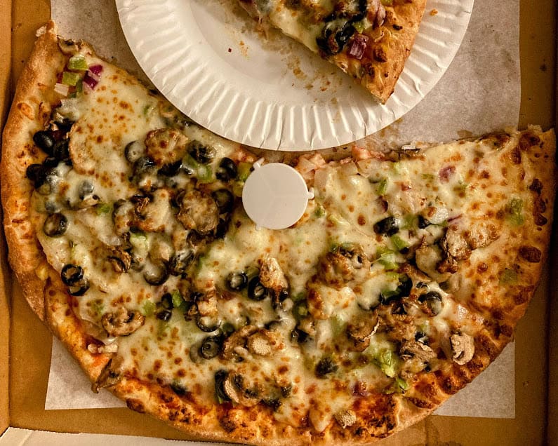 A close-up of a half-eaten large pizza loaded with mushrooms, olives, onions, and green peppers, placed in a takeout box with a white paper plate on top.