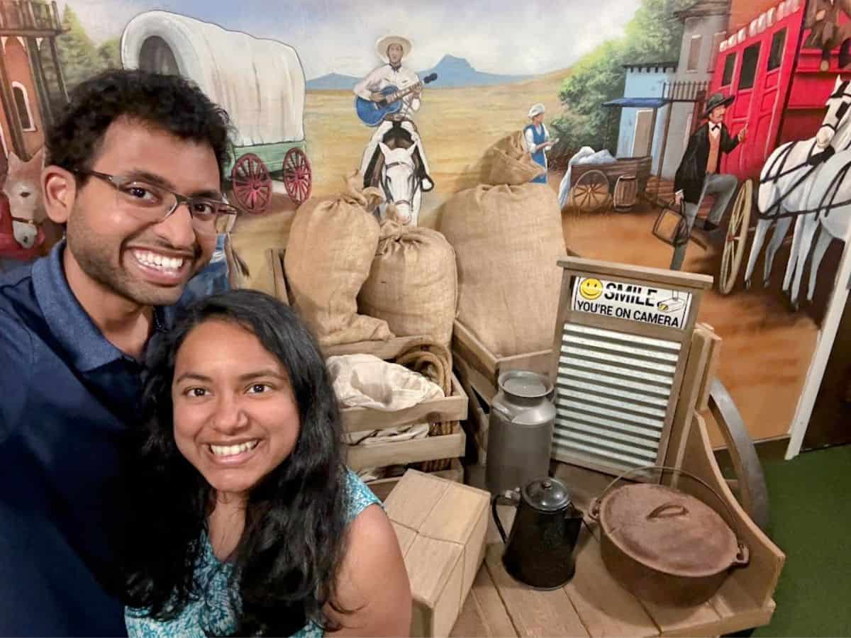 A cheerful couple poses in front of a colorful pioneer-themed backdrop with old-fashioned props, highlighting a fun photo spot mentioned in 32 Things To Do In Sacramento.