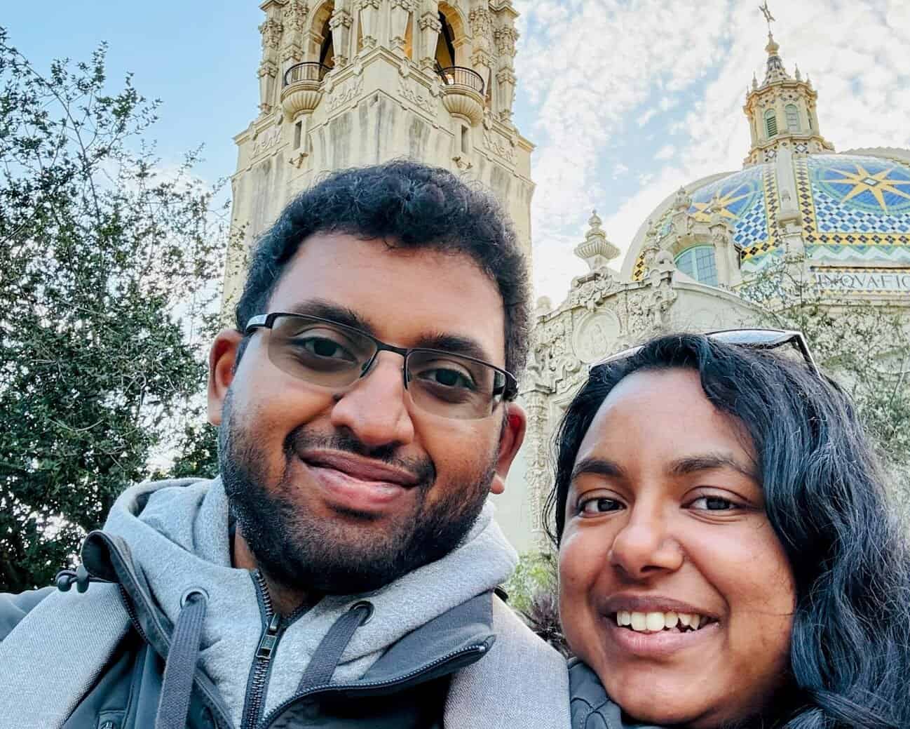 Kiran and Shreeyeh take a selfie in front of a historic building in San Diego, featuring an ornate tower and a colorful tiled dome under a partly cloudy sky.