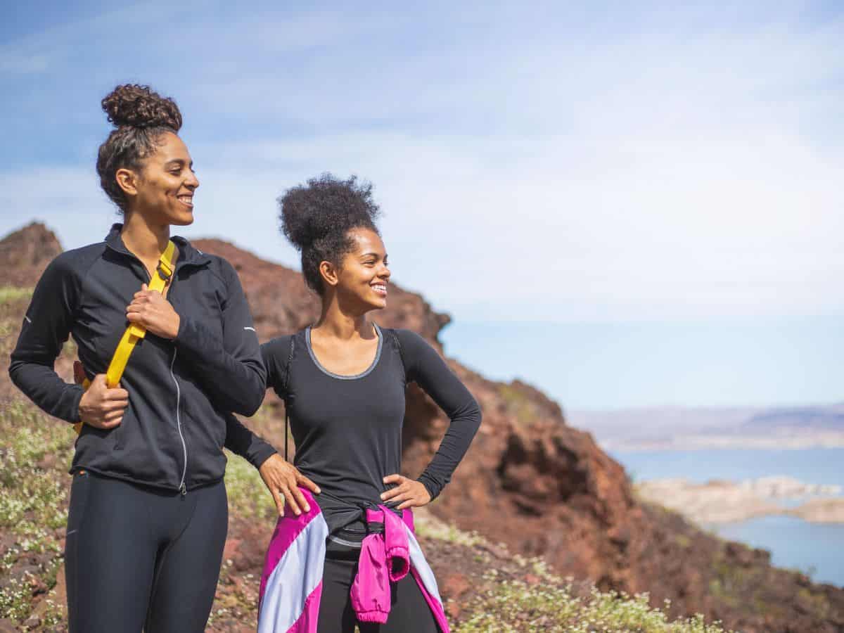 Two women in athletic wear smile while hiking a rocky trail with scenic views of a blue lake and distant mountains in the background.