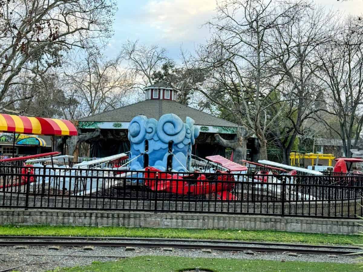 A small amusement ride with red and white airplanes surrounded by trees and a train track, part of the family-friendly fun featured in 32 Things To Do In Sacramento.