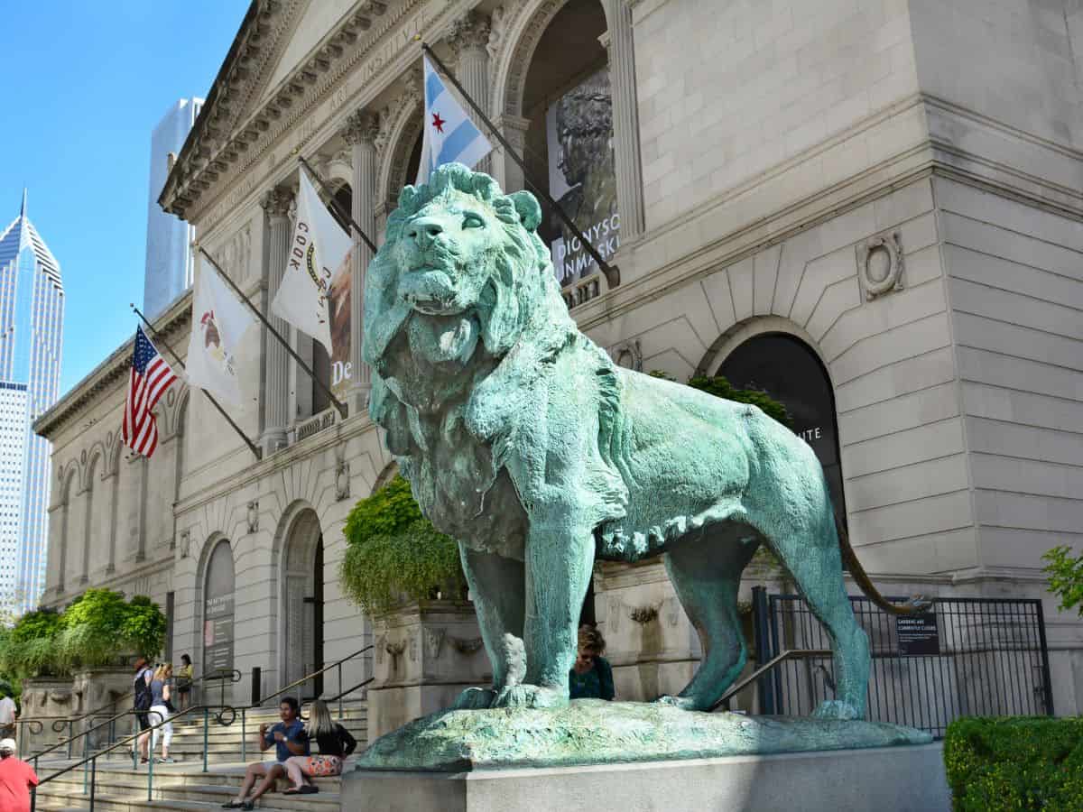 The iconic lion statue guarding the entrance of the Art Institute of Chicago with city flags and people on the steps—this world-class museum is one of the Top 10 Things to Do in Chicago, Ranked by Popularity.