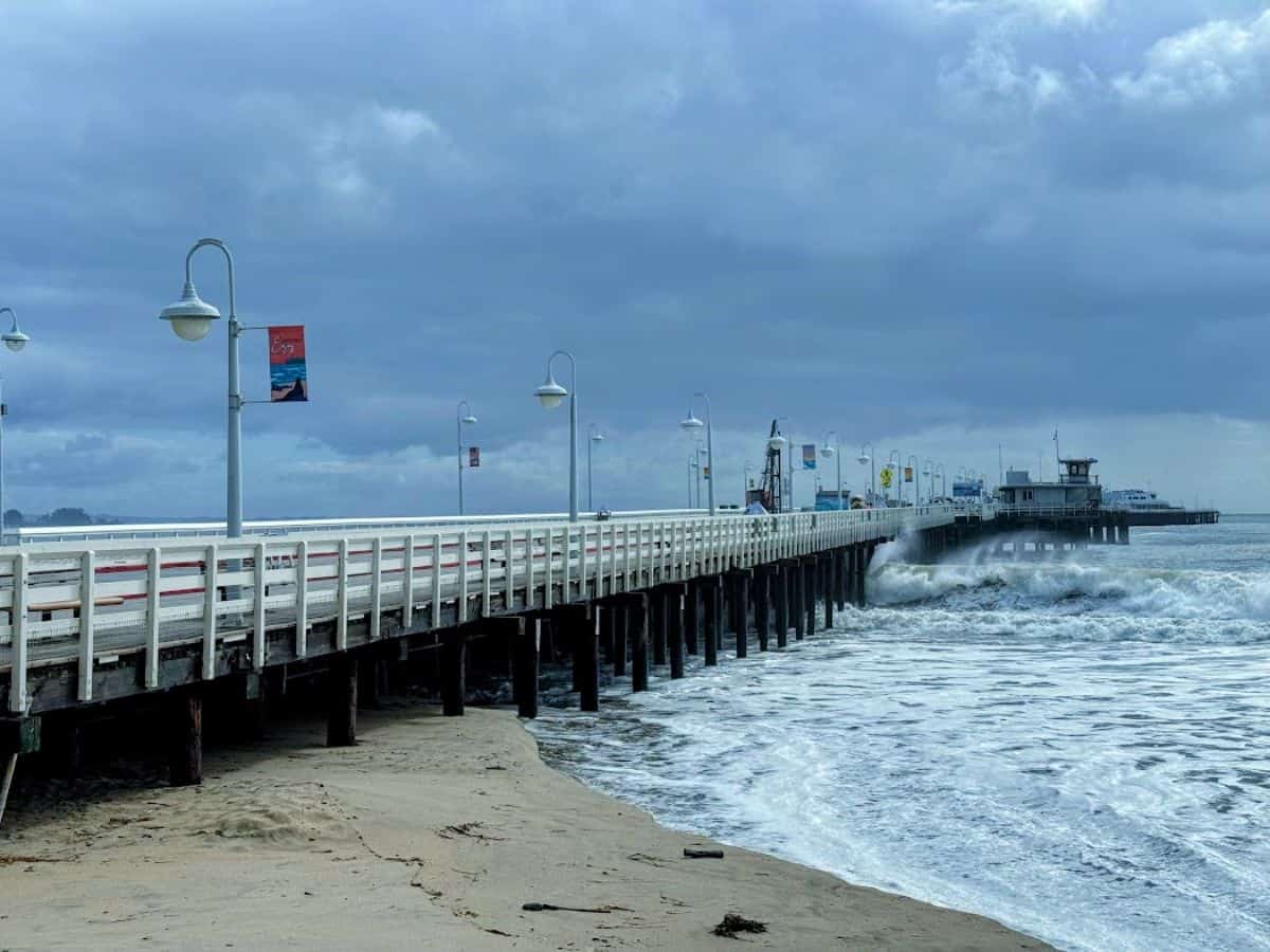 The Santa Cruz Wharf extends into the ocean, surrounded by waves under a cloudy sky. Dining on fresh seafood while enjoying oceanfront views is one of the 21 Things to Do in Santa Cruz.