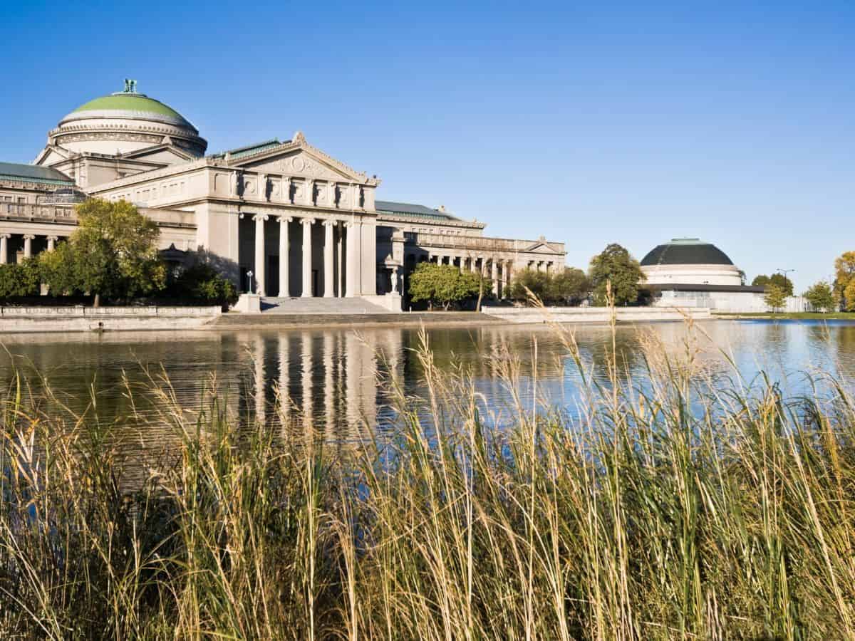 Exterior view of the grand Museum of Science and Industry surrounded by greenery and water, under a clear blue sky—exploring this massive museum is one of the Top 10 Things to Do in Chicago, Ranked by Popularity.