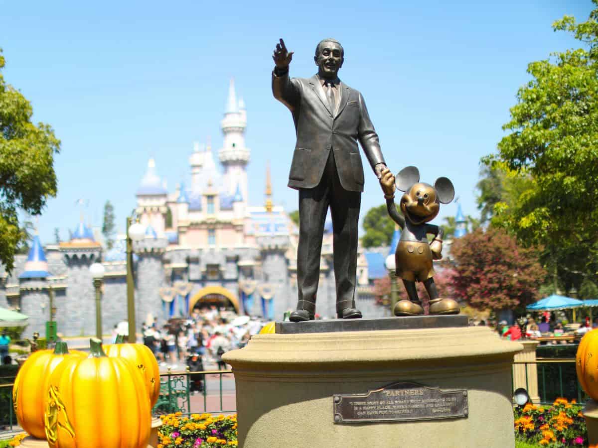 Bronze statue of Walt Disney holding Mickey Mouse’s hand in front of Sleeping Beauty Castle at Disneyland, surrounded by festive Halloween pumpkins and colorful flower beds.
