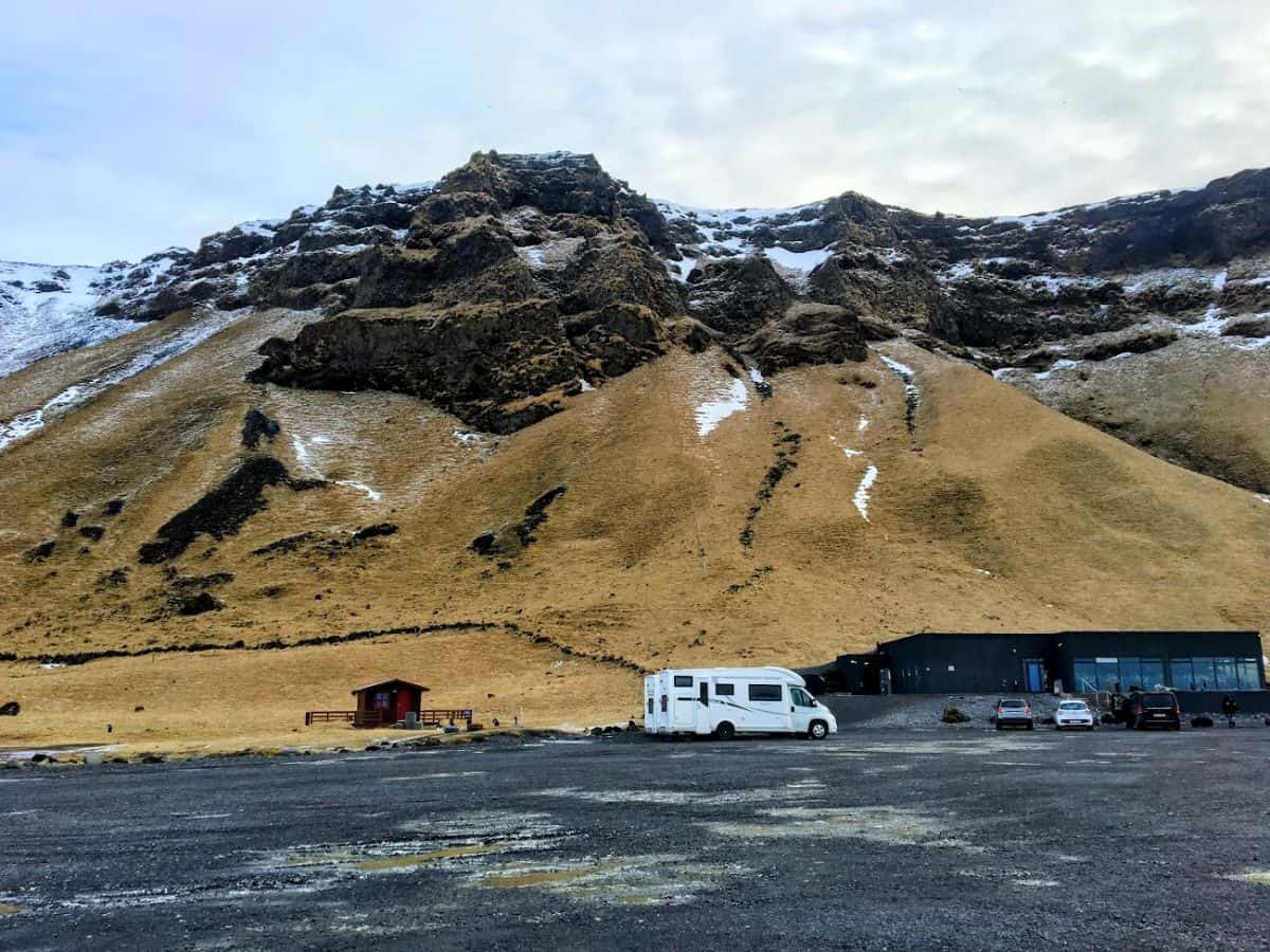 A camper van and a few cars are parked below a rugged cliff with streaks of snow and golden-brown moss. My Snowy Iceland Trip Was Equal Parts Stunning and Chaotic - Here's How to Be Ready for remote pit stops and dramatic landscapes.
