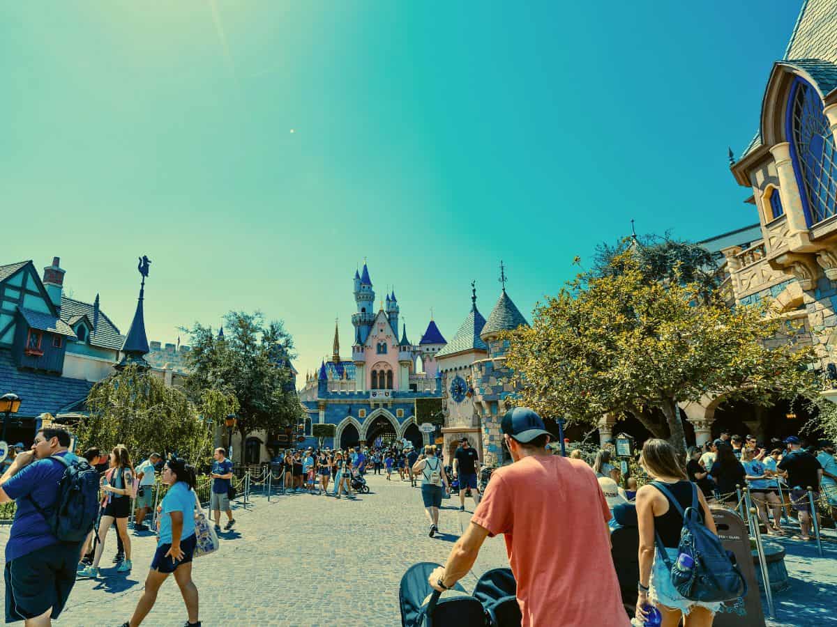 Visitors stroll through a bustling area of Disneyland near Sleeping Beauty Castle, surrounded by medieval-style buildings and trees, under a clear, sunny sky. The castle's iconic blue spires rise in the distance.