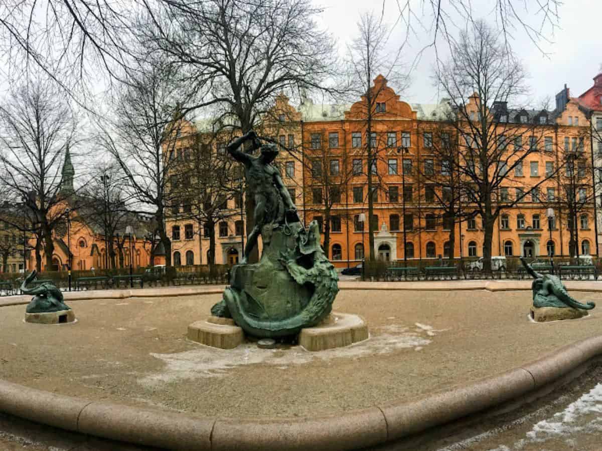 A dramatic bronze statue of a man slaying a dragon stands in the center of Mariatorget, a quiet city square surrounded by bare trees and ornate peach-toned buildings in winter.