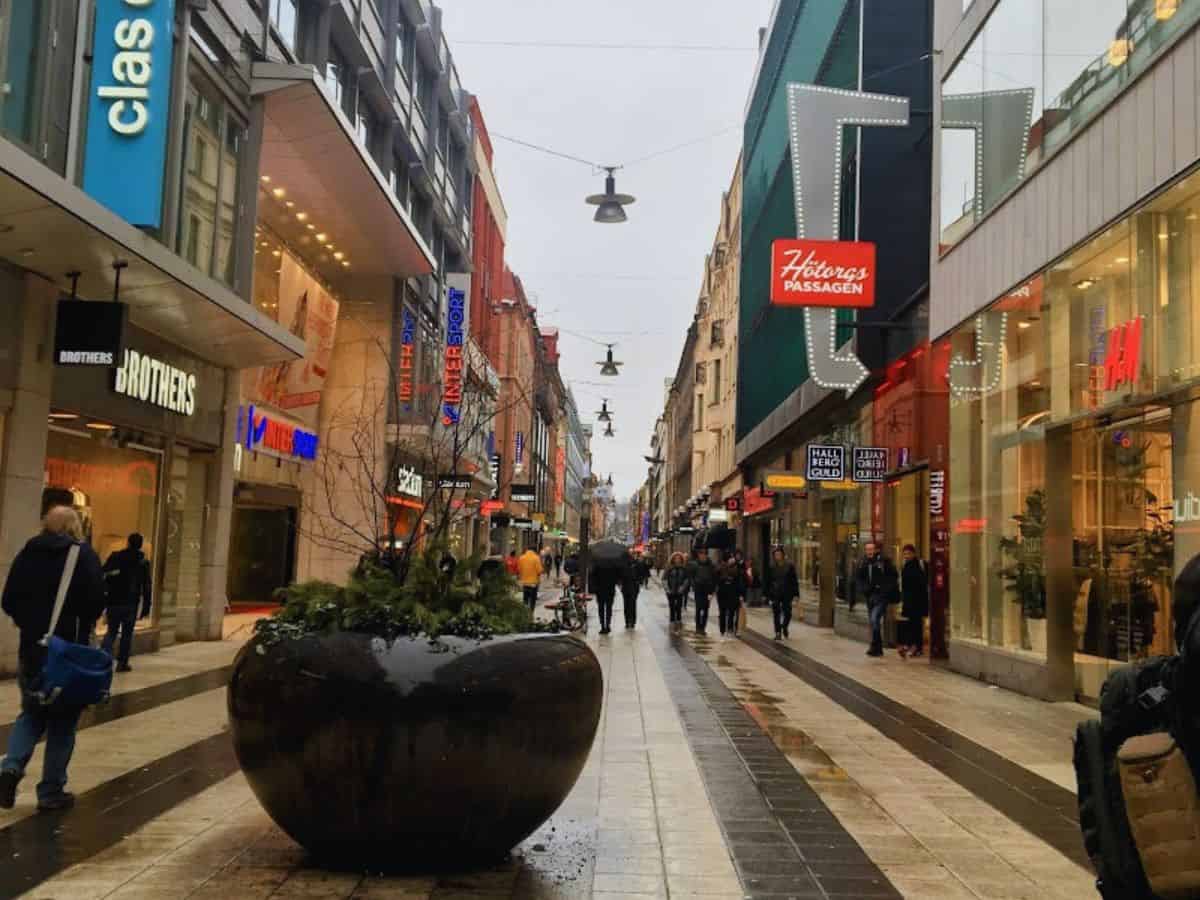 People stroll along Drottninggatan, a bustling pedestrian shopping street in Stockholm lined with big-name stores like H&M and Intersport, beneath gray skies and glowing signs.