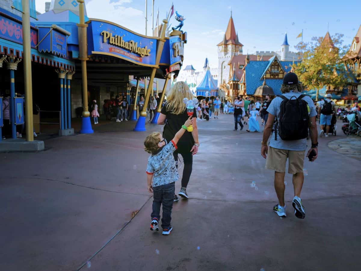 A young child holding a Mickey Mouse bubble wand walks hand-in-hand with a woman through a whimsical Fantasyland area in Disneyland, passing the PhilharMagic attraction surrounded by other families and castle-like buildings.