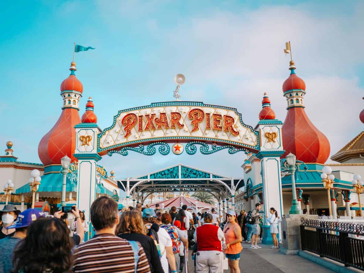 A crowd of parkgoers walks under the colorful Pixar Pier entrance arch at Disney California Adventure, framed by bright red turrets and detailed architectural designs on a sunny day.
