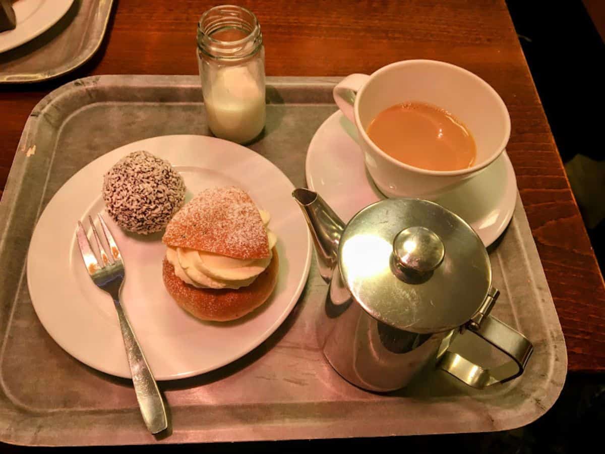 A traditional Swedish fika spread with a semla (cream bun), chokladboll, coffee with milk, and a silver teapot served on a cafeteria tray in a cozy café.