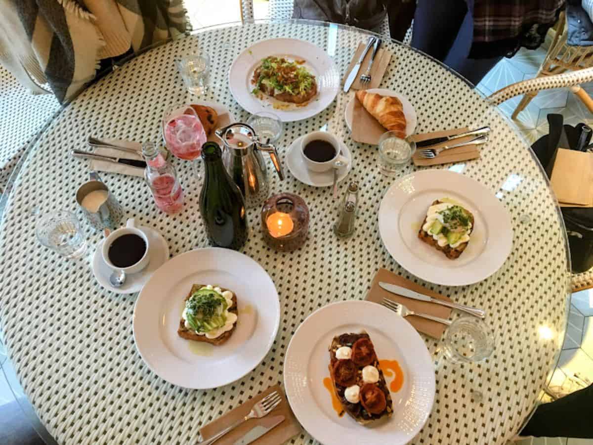 A chic brunch spread at Greta’s Café featuring beautifully plated open-faced sandwiches topped with avocado and greens, pastries, and coffee, all on a white wicker table with cozy vibes.