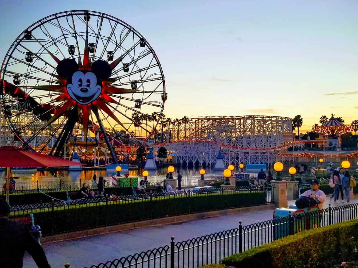 Sunset view of Pixar Pal-A-Round Ferris wheel with Mickey Mouse’s face in the center, set beside roller coasters and carnival lights at Disney California Adventure.