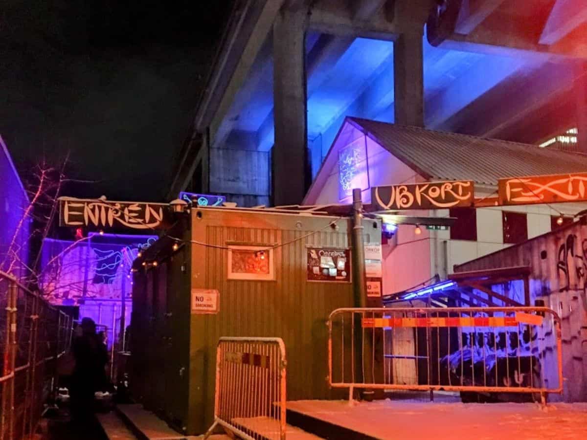A gritty, neon-lit entrance to the Under Bron nightclub tucked beneath a highway overpass, with graffiti-style signs, metal barriers, and a cool urban underground vibe.