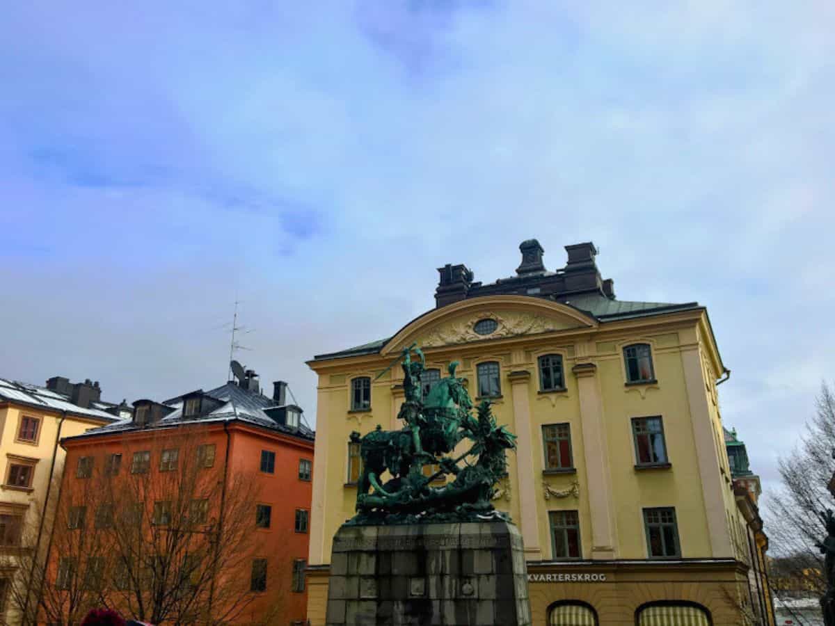 A dramatic bronze statue of St. George slaying a dragon stands in a small square in Gamla Stan, Stockholm, surrounded by colorful historic buildings under a partly cloudy sky.