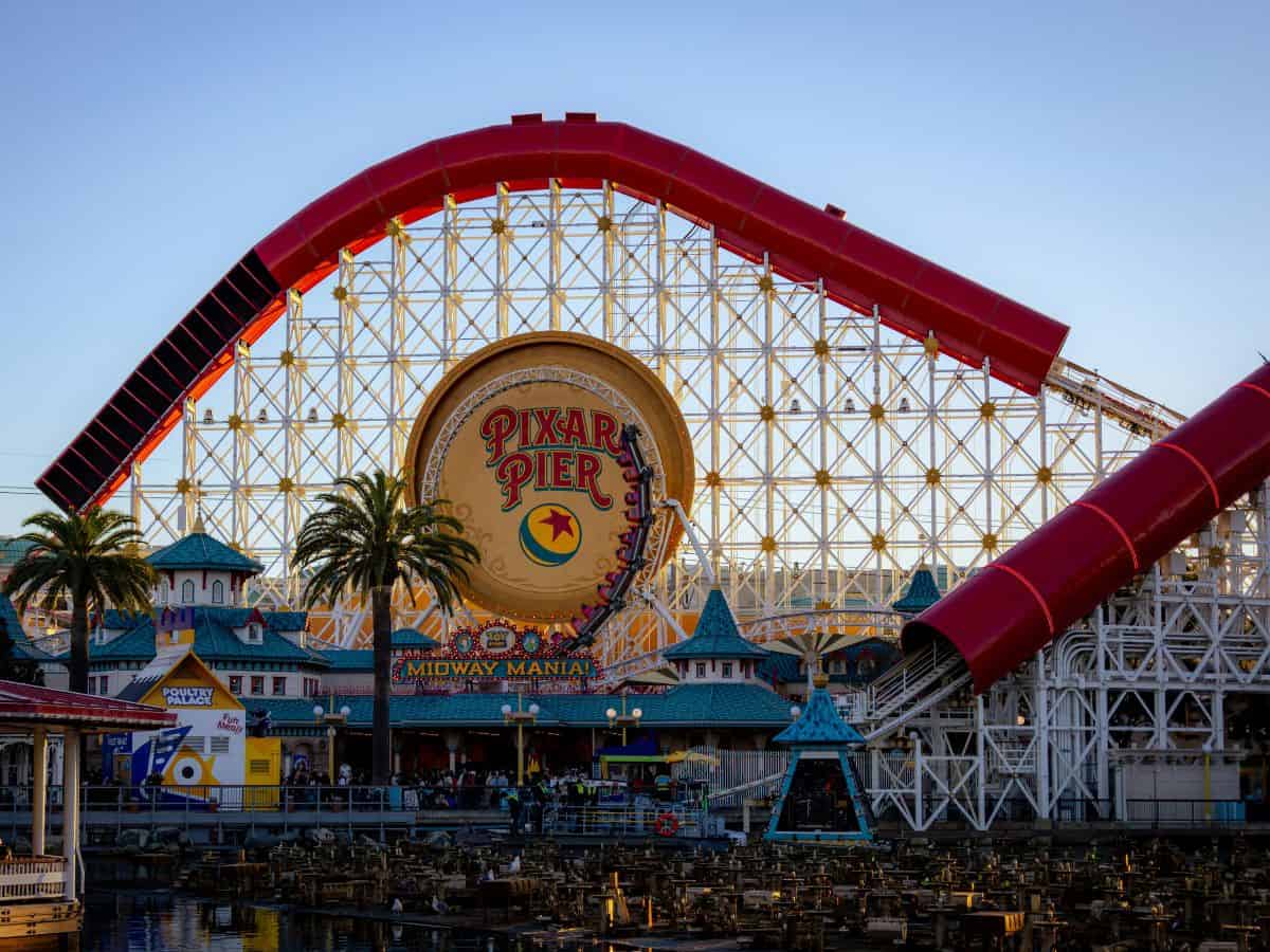 Pixar Pier at California Adventure with the massive red roller coaster loop arching over the Pixar Pier sign, flanked by palm trees and whimsical carnival-style buildings.