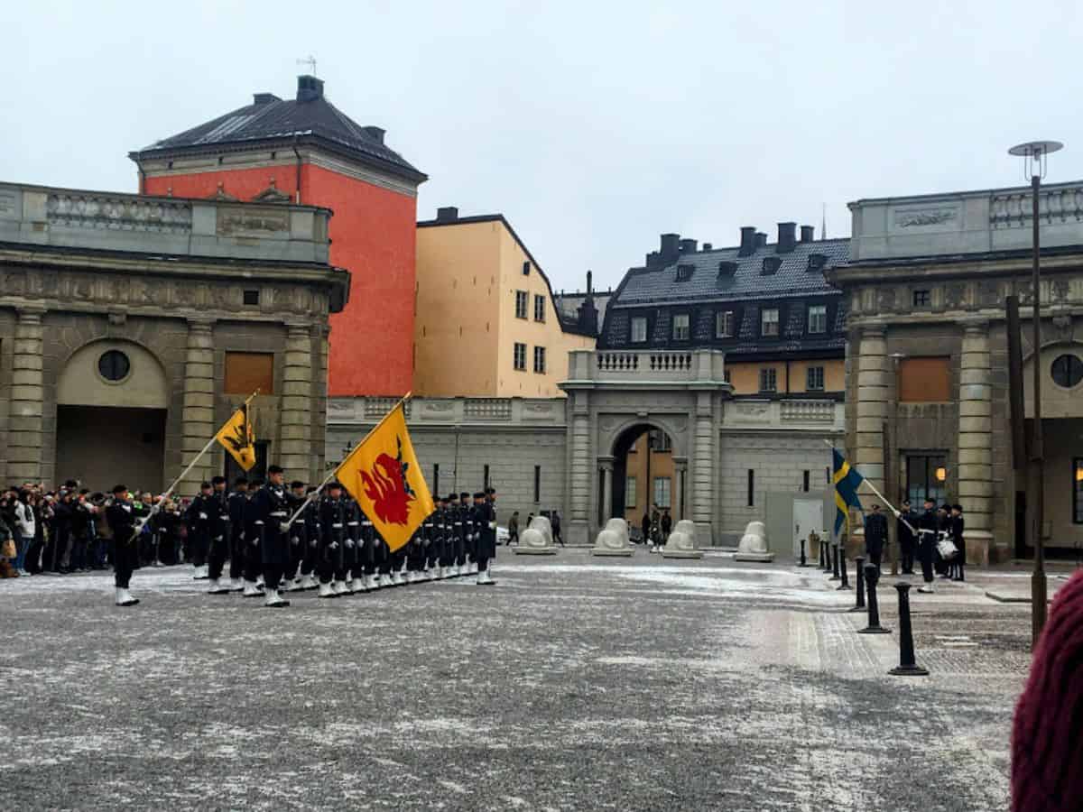Uniformed guards in formation perform the ceremonial Changing of the Guard in front of a historic Stockholm building, with flags and a gathered crowd watching.