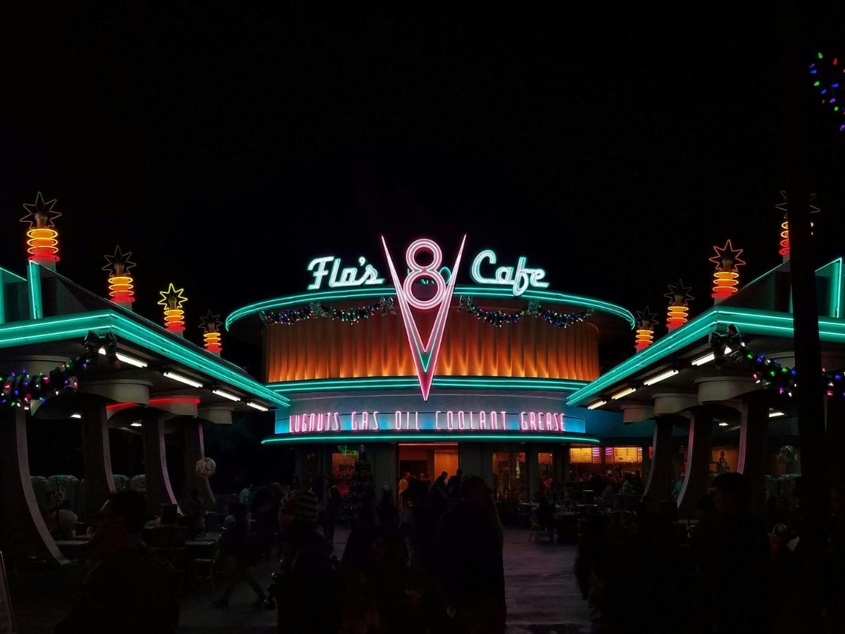 Flo’s V8 Cafe glowing in neon lights at night in Cars Land, styled as a retro diner with teal, pink, and orange accents and decorated with holiday garlands.