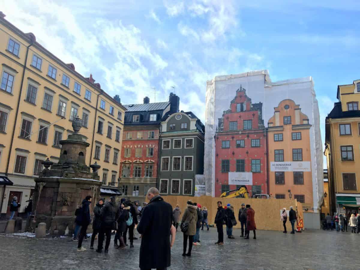 Tourists mill about Stortorget, Stockholm’s oldest square, where iconic red and yellow townhouses are partly wrapped in construction tarps printed to mimic their facades.