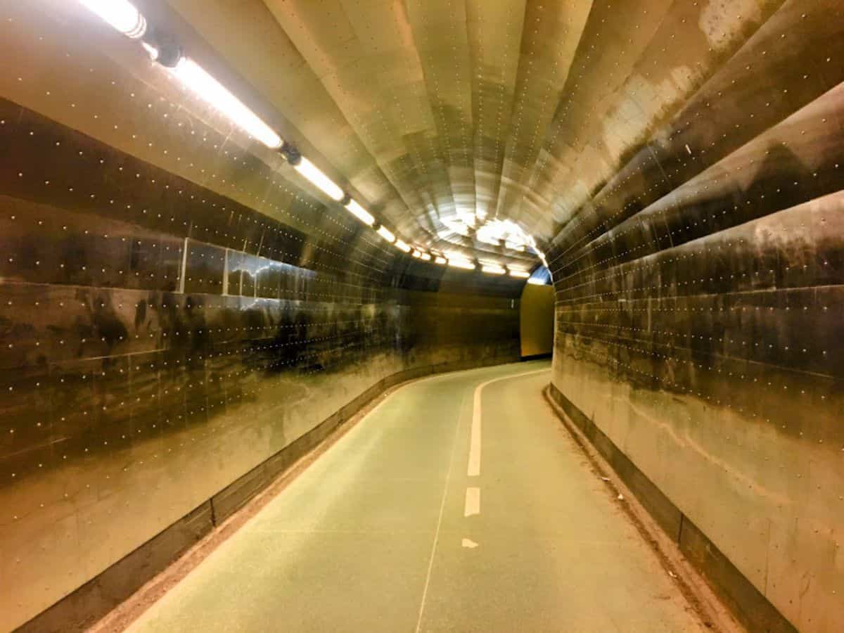 A softly lit, metallic pedestrian tunnel curves ahead with a faint green path line, creating a futuristic and moody atmosphere.