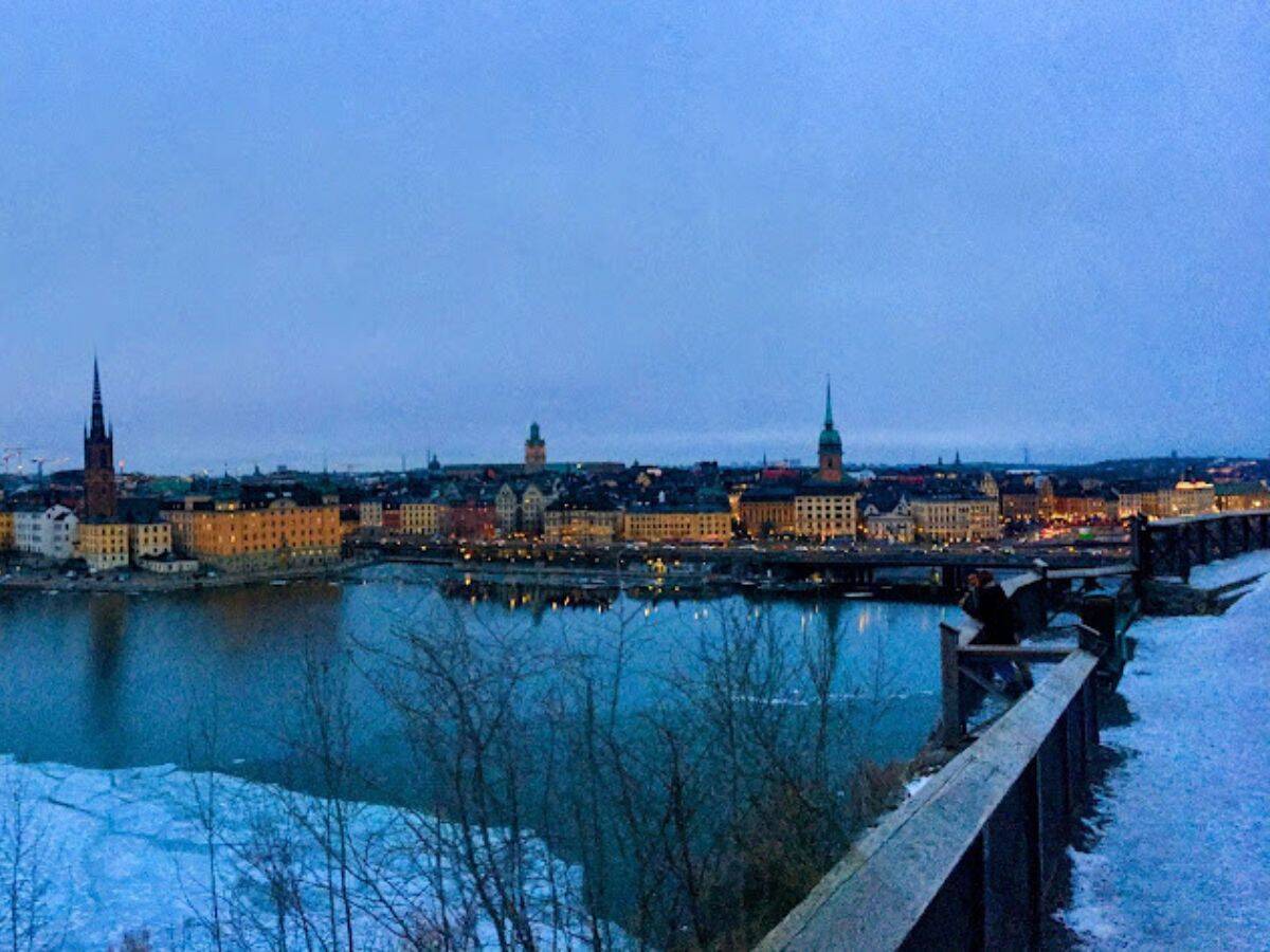 Panoramic view of central Stockholm from Mariaberget, with icy waters below, historic buildings, church spires, and twinkling evening lights stretching into the distanc