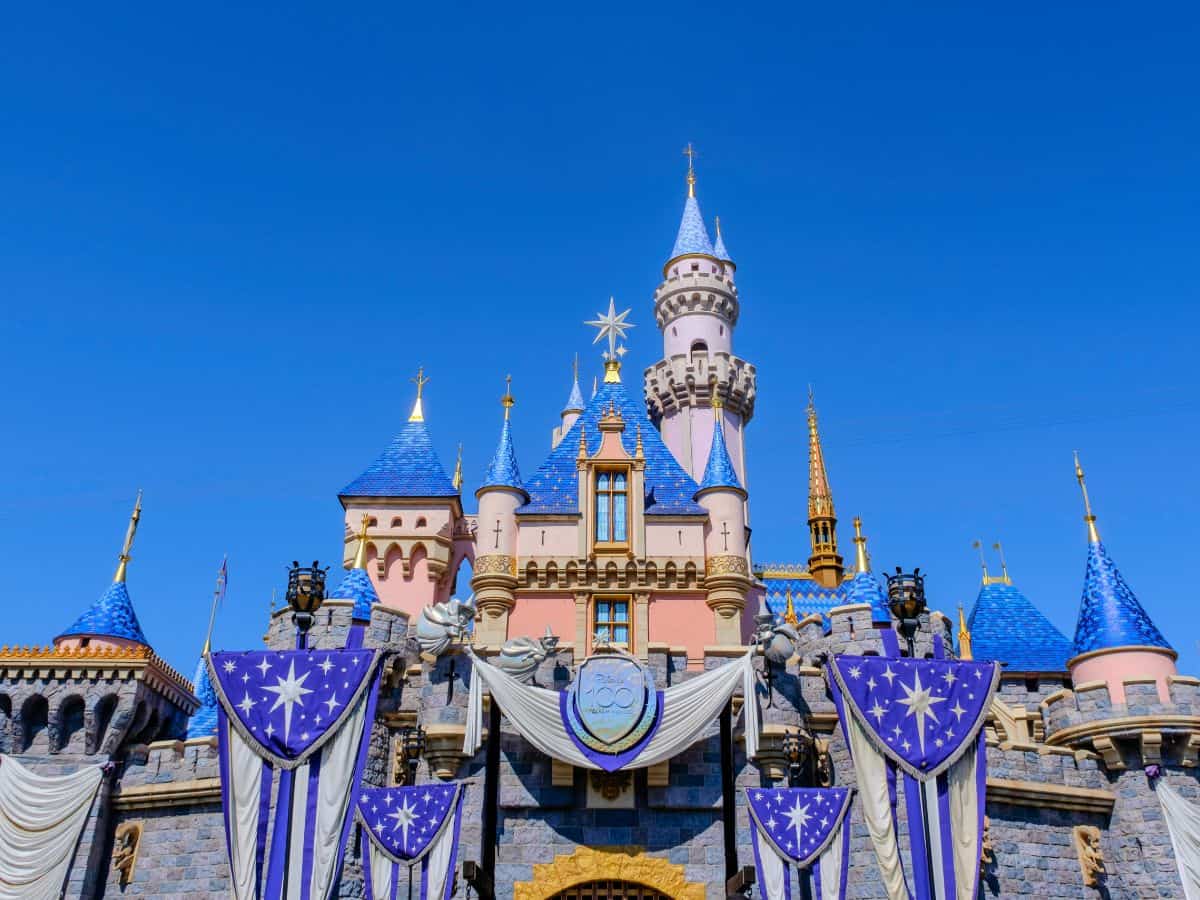 Front view of Disneyland’s Sleeping Beauty Castle decorated for the Disney 100 celebration with royal blue banners, silver stars, and sparkling spires.