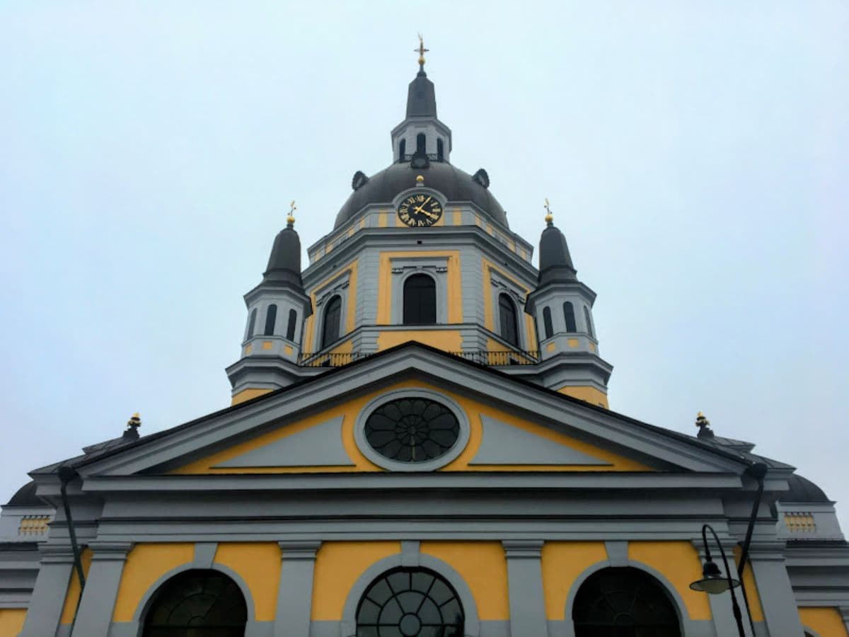 The pale yellow and gray exterior of Katarina Kyrka, a baroque-style church with a domed clock tower and smaller spires, rises against a cloudy sky.