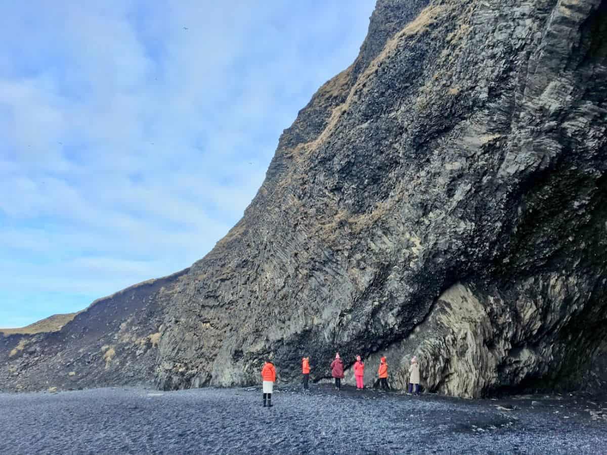 A group of people in colorful jackets stands at the base of a massive cliff wall on a black sand beach. My Snowy Iceland Trip Was Equal Parts Stunning and Chaotic - Here's How to Be Ready to feel tiny next to Iceland's epic natural sculptures.