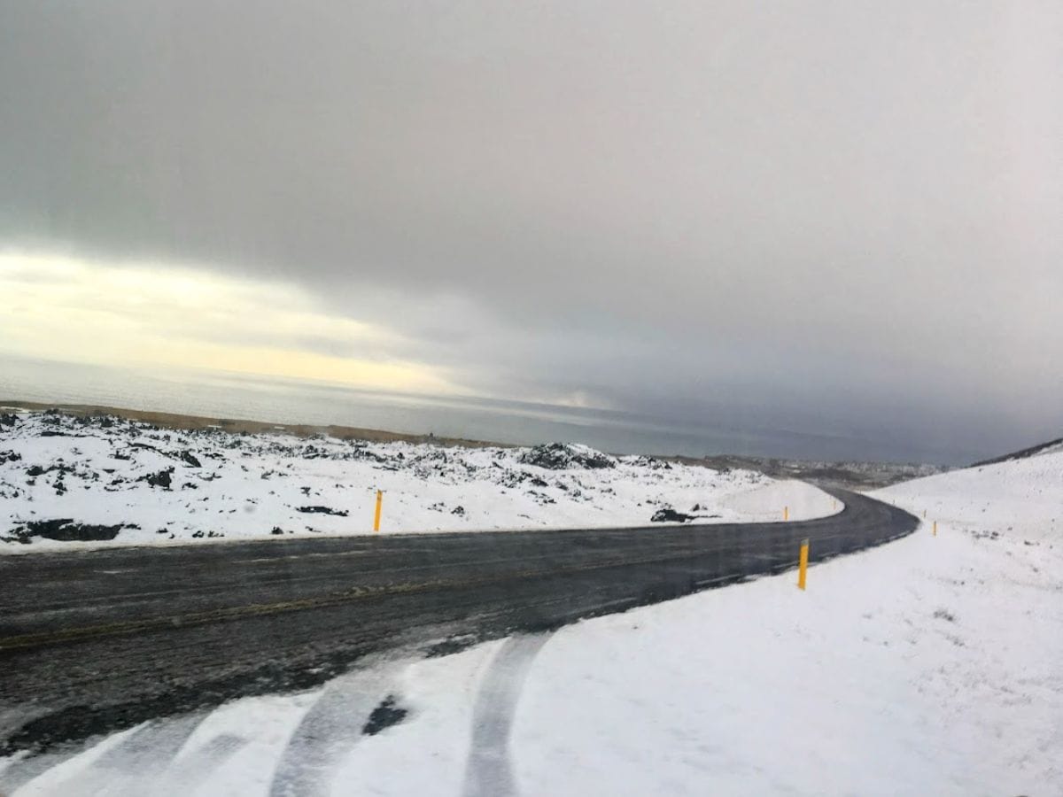 A snow-dusted road winds toward the ocean under a heavy gray sky, flanked by dark lava rocks. My Snowy Iceland Trip Was Equal Parts Stunning and Chaotic - Here's How to Be Ready for slippery curves and sudden weather shifts.