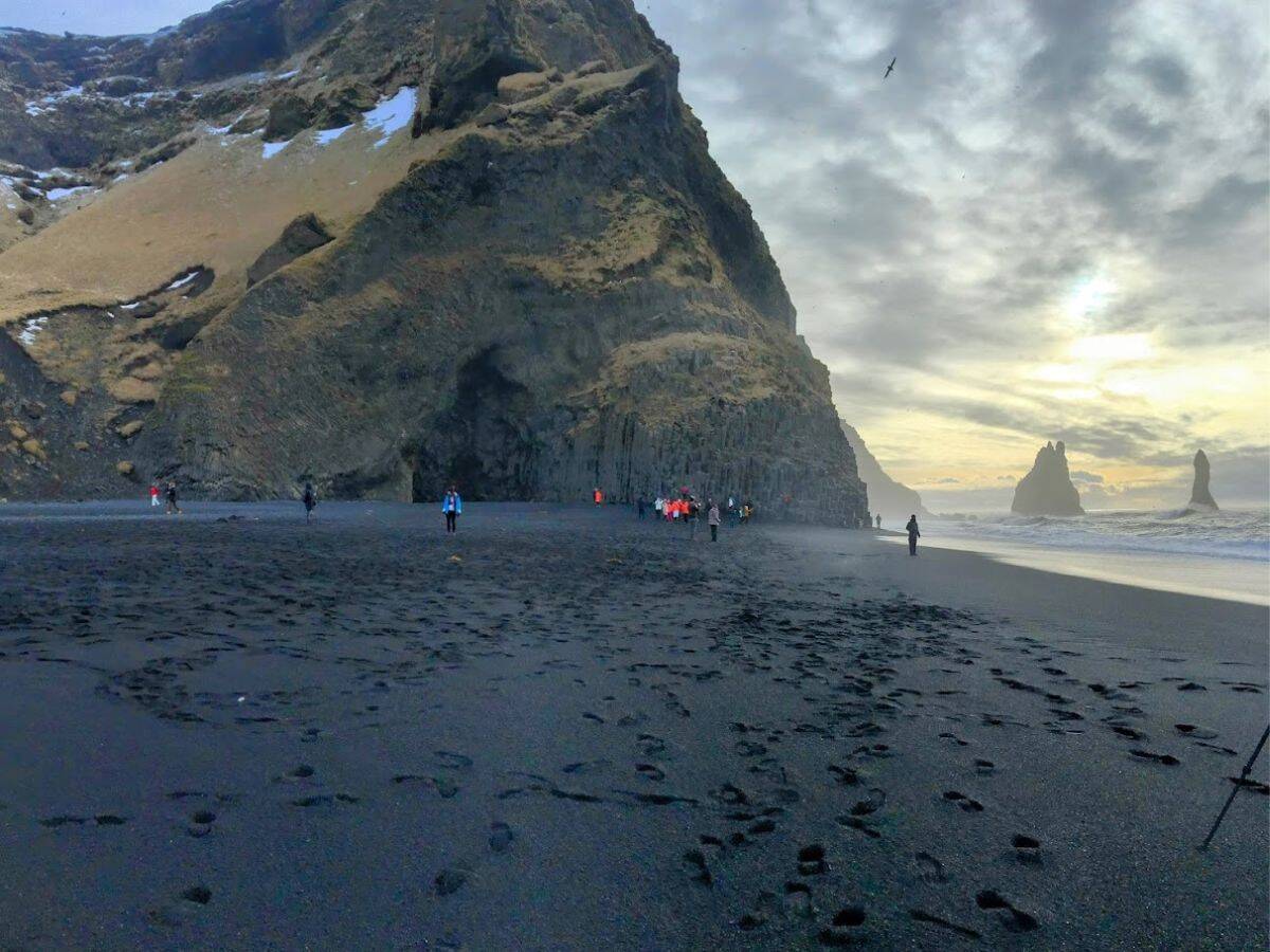 Travelers explore Iceland’s famous black sand beach with towering cliffs and sea stacks under a moody sky. My Snowy Iceland Trip Was Equal Parts Stunning and Chaotic - Here's How to Be Ready for wind, waves, and surreal beauty.