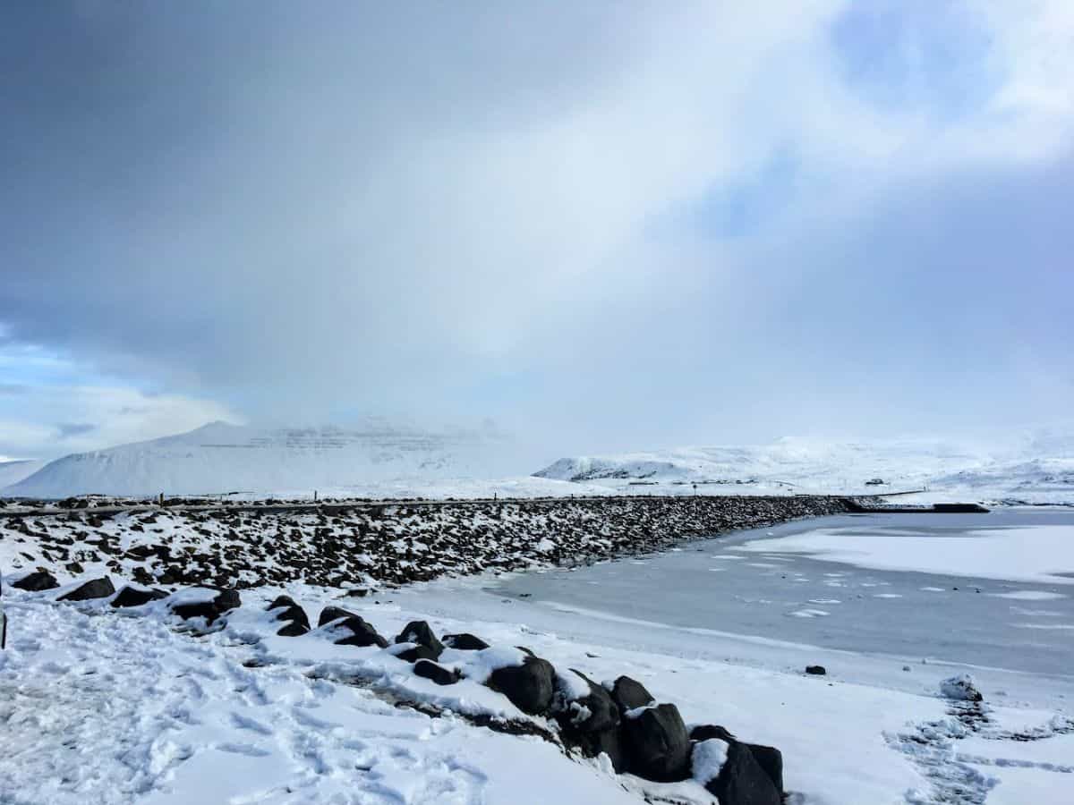 A snowy coastal scene with icy water and distant white-capped mountains shrouded in clouds. My Snowy Iceland Trip Was Equal Parts Stunning and Chaotic - Here's How to Be Ready for unpredictable winter weather and mesmerizing views.