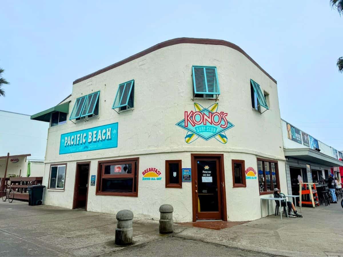 Corner view of Kono’s Surf Club Café in Pacific Beach, San Diego, a sandy-colored building with teal shutters and signage that reads “Pacific Beach” and “Kono’s Surf Club.” A sign near the entrance highlights “Breakfast served all day,” and a few people are seated at outdoor tables along the sidewalk.