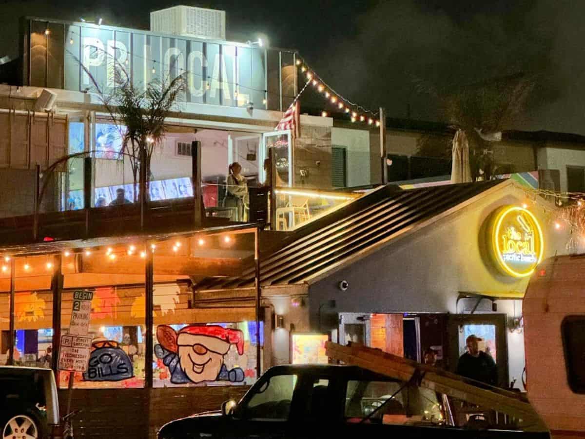 A bustling night scene at 'The Local,' highlighted by its brightly lit sign and a glowing rooftop sign spelling out "PB LOCAL" in large letters. Patrons can be seen in the upper-level outdoor seating, making it a lively addition to the best bars in Pacific Beach San Diego
