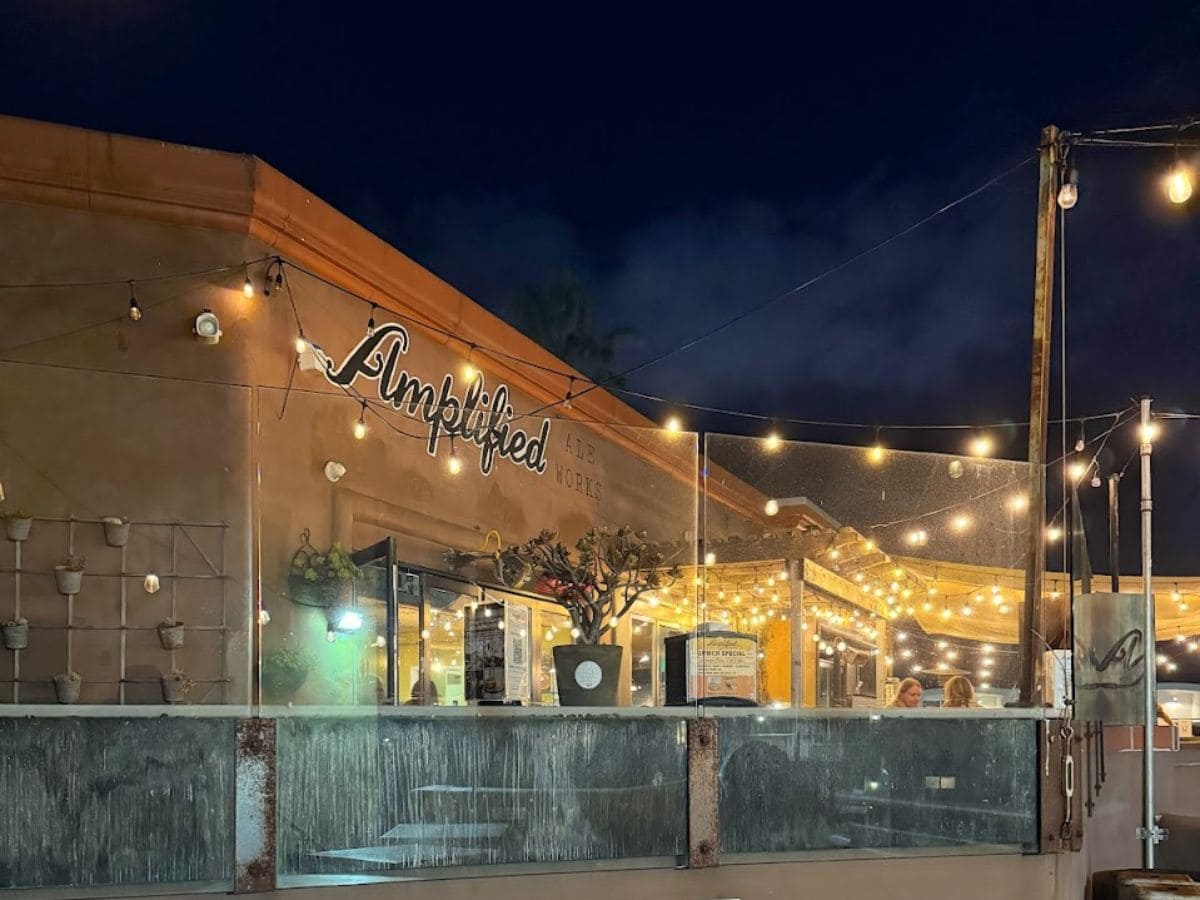 The entrance to 'Amplified Ale Works' is marked by a wooden sign with playful lettering, pointing to a warmly lit beer garden. This photo captures the welcoming atmosphere of the bar, a gem among the best bars in Pacific Beach San Diego, with its promise of good times just an arrow sign away
