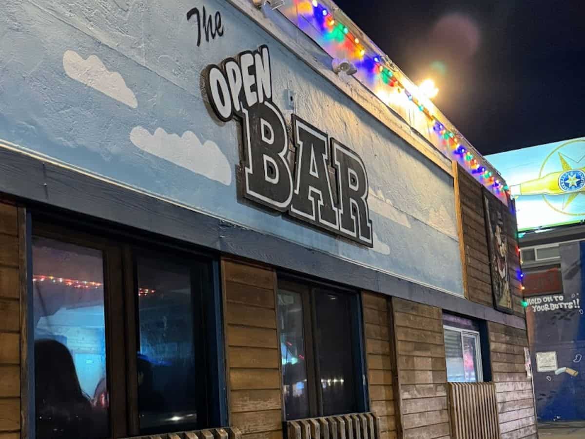 This image shows 'The Open Bar,' one of the best bars in Pacific Beach San Diego, at night. The establishment's name is displayed in large, cut-out letters mounted on a wall with a painted illusion of blue sky and fluffy clouds, accompanied by festive, multicolored string lights enhancing the inviting ambiance.