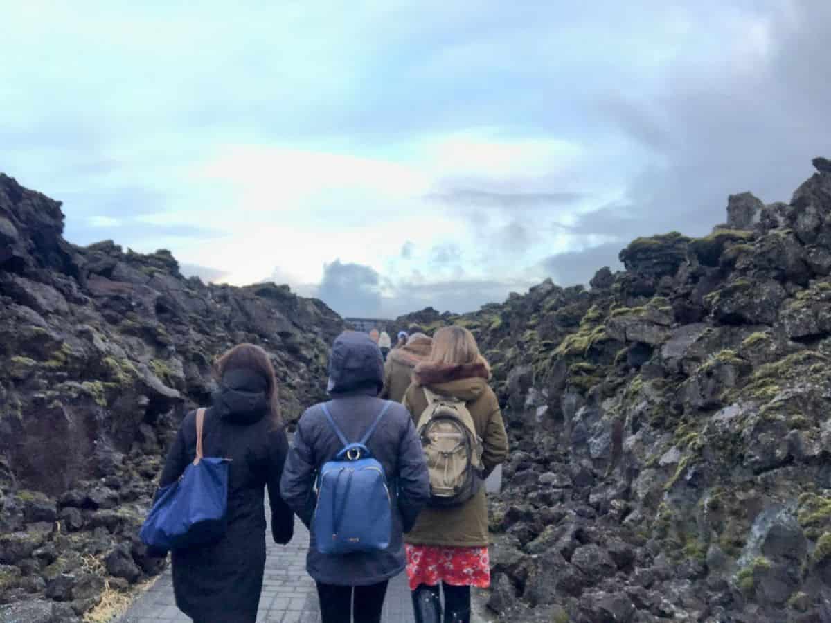 A group of travelers in winter jackets walk through a narrow lava field path under a moody sky. My Snowy Iceland Trip Was Equal Parts Stunning and Chaotic - Here's How to Be Ready for chilly treks through Iceland’s dramatic geology.
