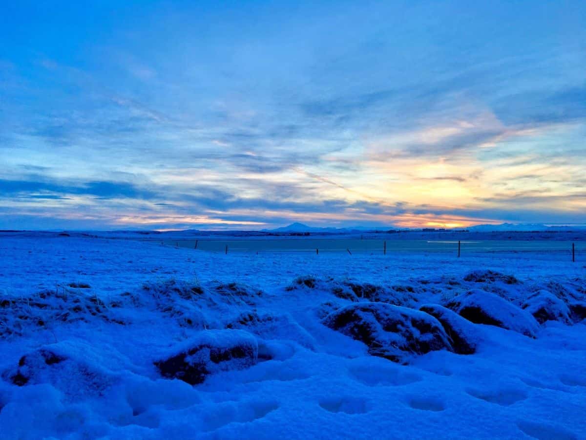 A vivid blue and orange sunset casts warm light over a snowy field and distant mountains. My Snowy Iceland Trip Was Equal Parts Stunning and Chaotic - Here's How to Be Ready for magical light at unexpected times.