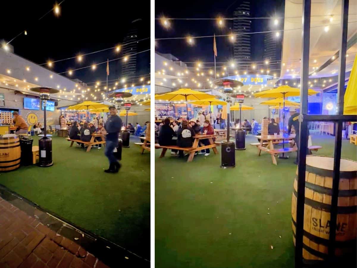 Outdoor beer garden at Happy Does bar, with yellow umbrellas over picnic tables, twinkling string lights above, and patrons enjoying drinks and games in a lively nighttime setting.