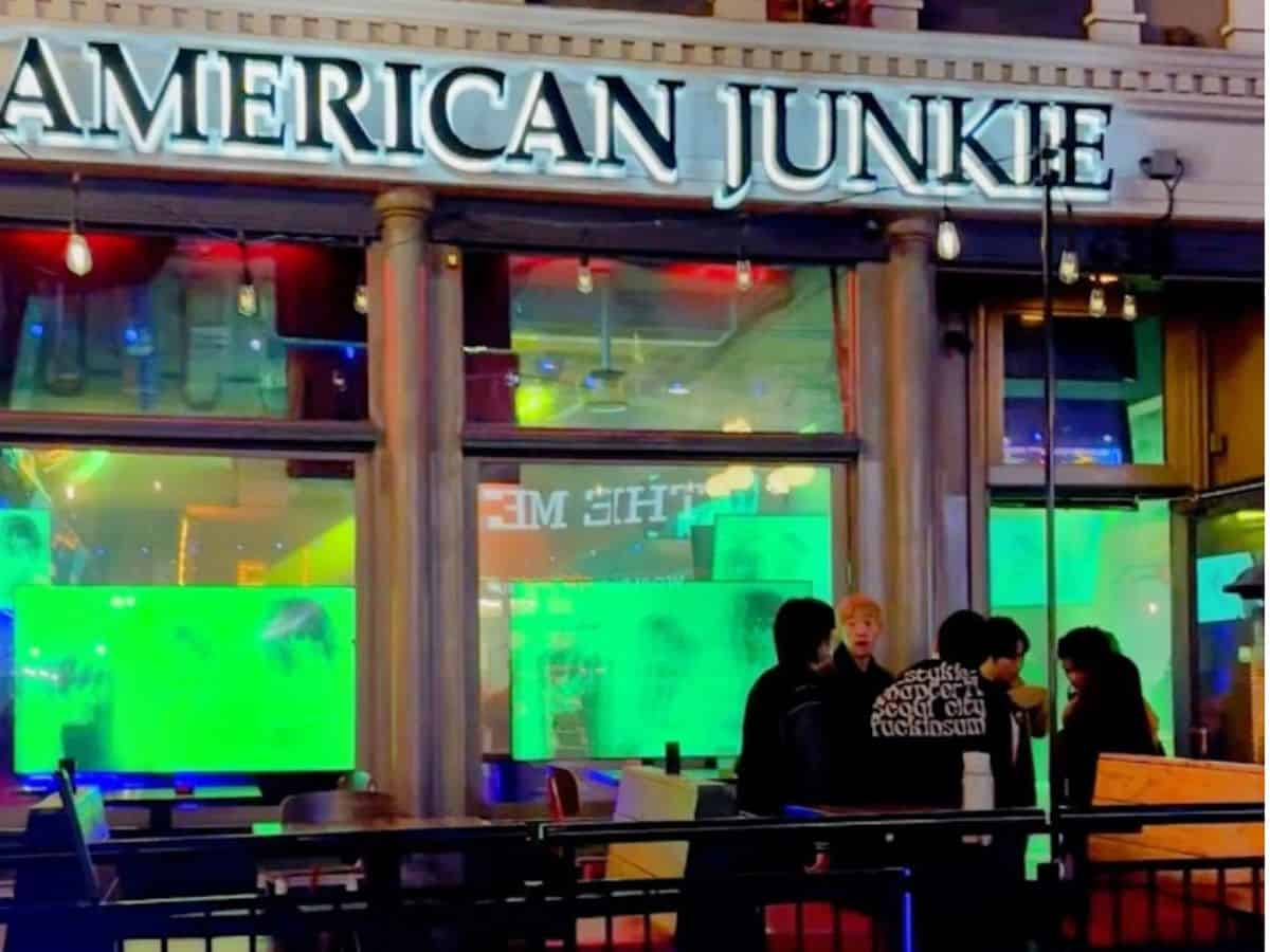 Front of American Junkie, a bar with neon-lit windows glowing green and red. A group of people gathers outside the entrance under the bar's glowing sign.