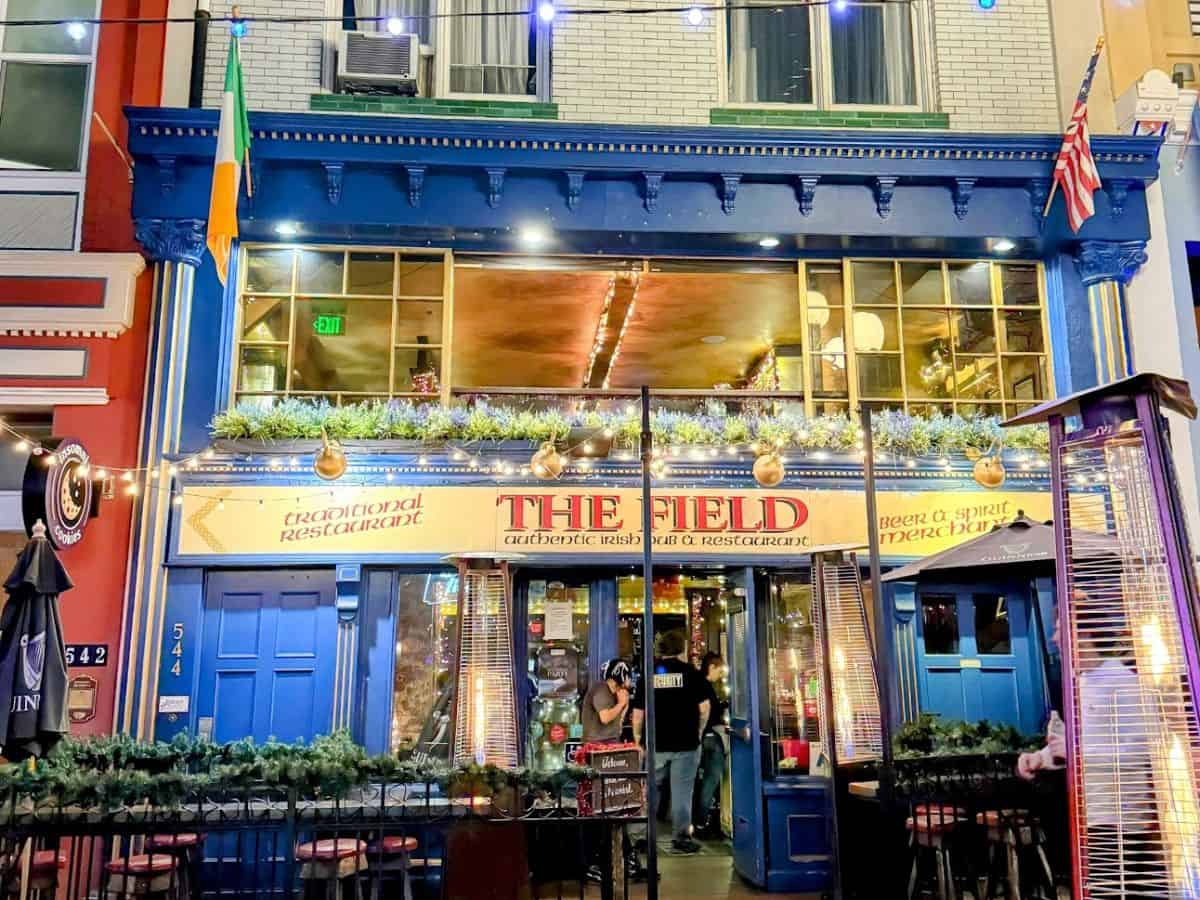 The colorful facade of The Field Irish Pub in San Diego's Gaslamp Quarter, decorated with garlands and lights for the holidays. The warm glow inside spills out to the sidewalk seating area.