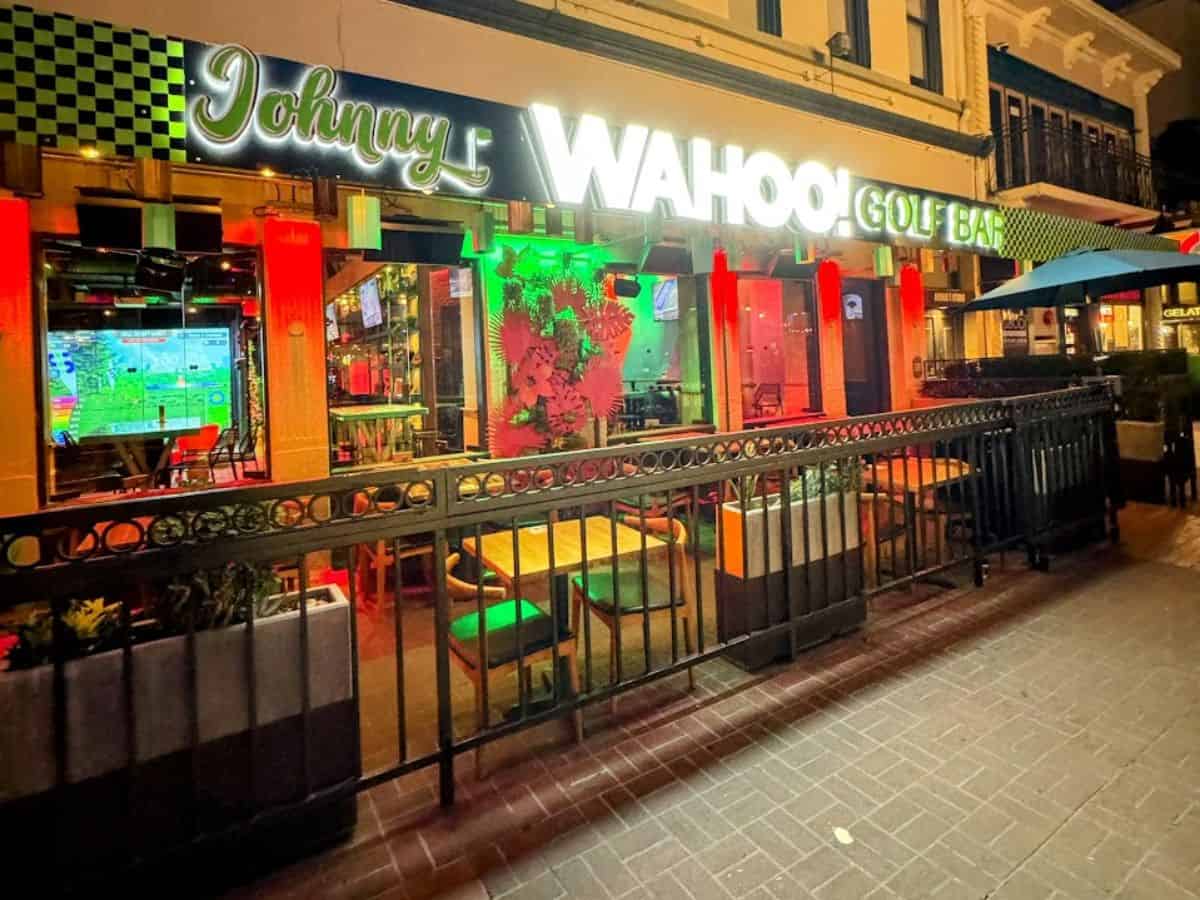 Nighttime view of Swing Social, a lively outdoor bar and golf-themed venue featuring string lights, wooden picnic tables, barrels, and large digital golf simulators inside. Patrons are gathered under yellow umbrellas.