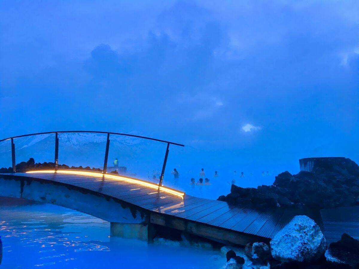A glowing footbridge stretches over steaming blue waters at the Blue Lagoon, with snowy mountains in the backdrop. My Snowy Iceland Trip Was Equal Parts Stunning and Chaotic - Here's How to Be Ready to soak off the chill in iconic geothermal waters.