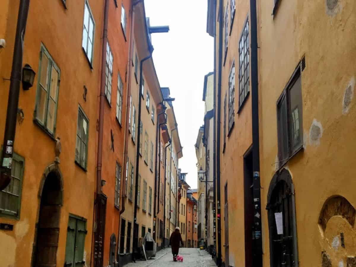 A cobblestone alley in Stockholm's Old Town, Gamla Stan, lined with tall, colorful buildings in earthy yellows and oranges, with a person walking in the distance.