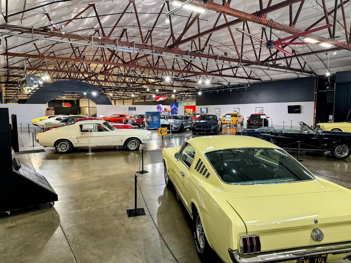 A spacious indoor exhibit at the California Automobile Museum in Sacramento featuring a lineup of classic cars, including multiple vintage Mustangs, under a warehouse-style roof with exposed beams.