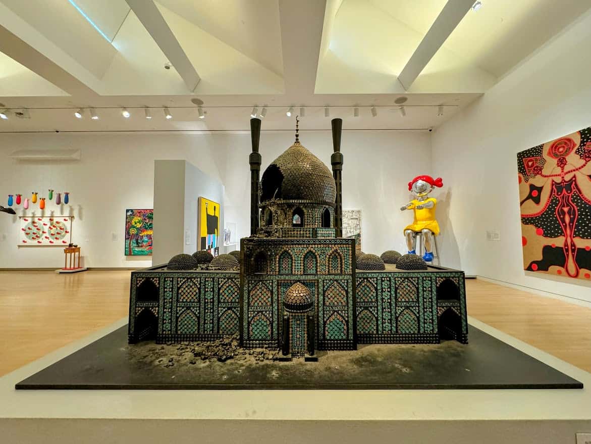 An eclectic gallery inside the Crocker Art Museum showcasing colorful modern art, including a detailed miniature mosque model in the foreground and whimsical sculptures and abstract paintings in the background.