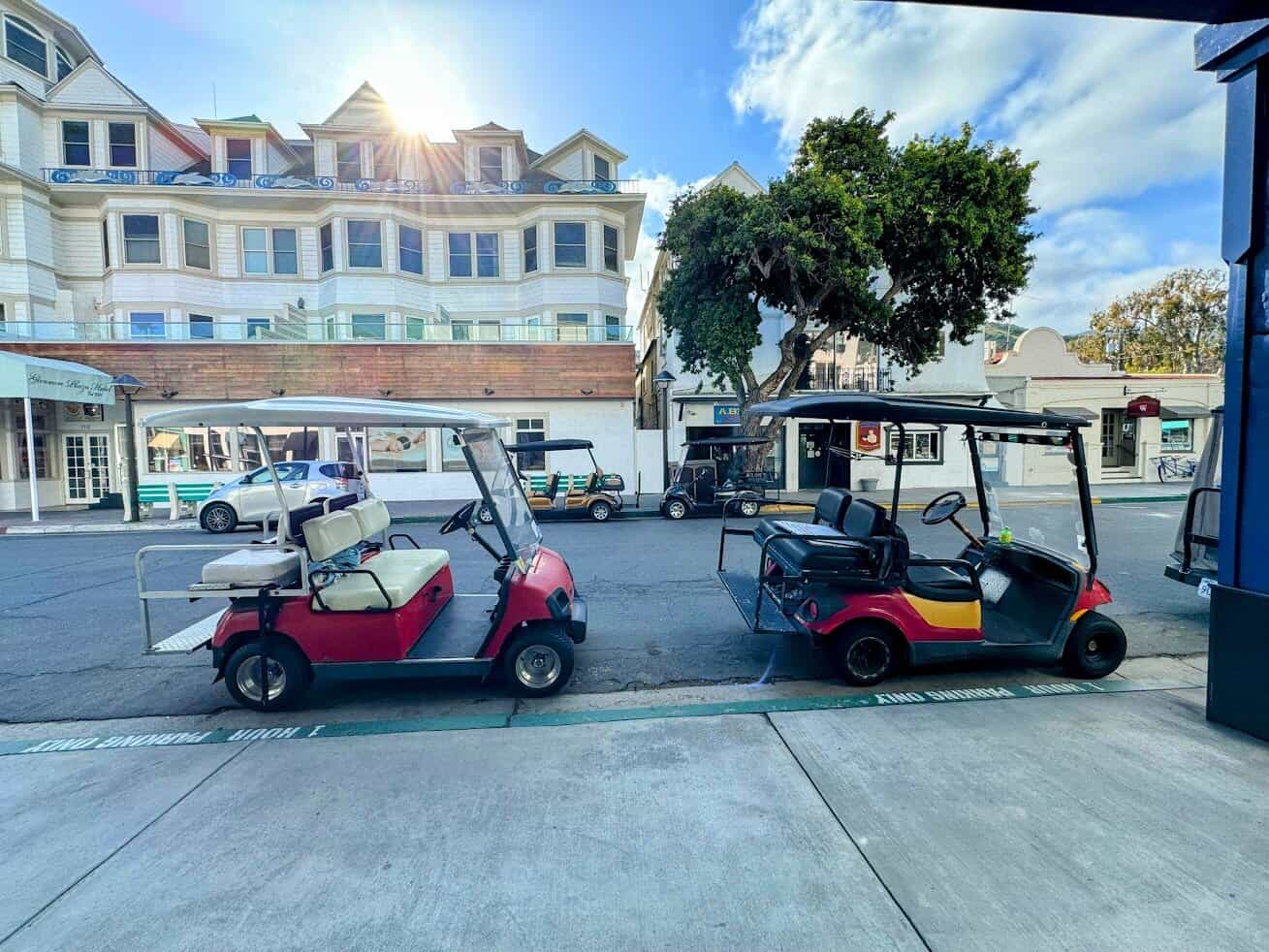 Two golf carts, one red and one yellow-black, are parked on a street in Avalon, Catalina Island, with a large Victorian-style building and the sun peeking over the rooftop.

