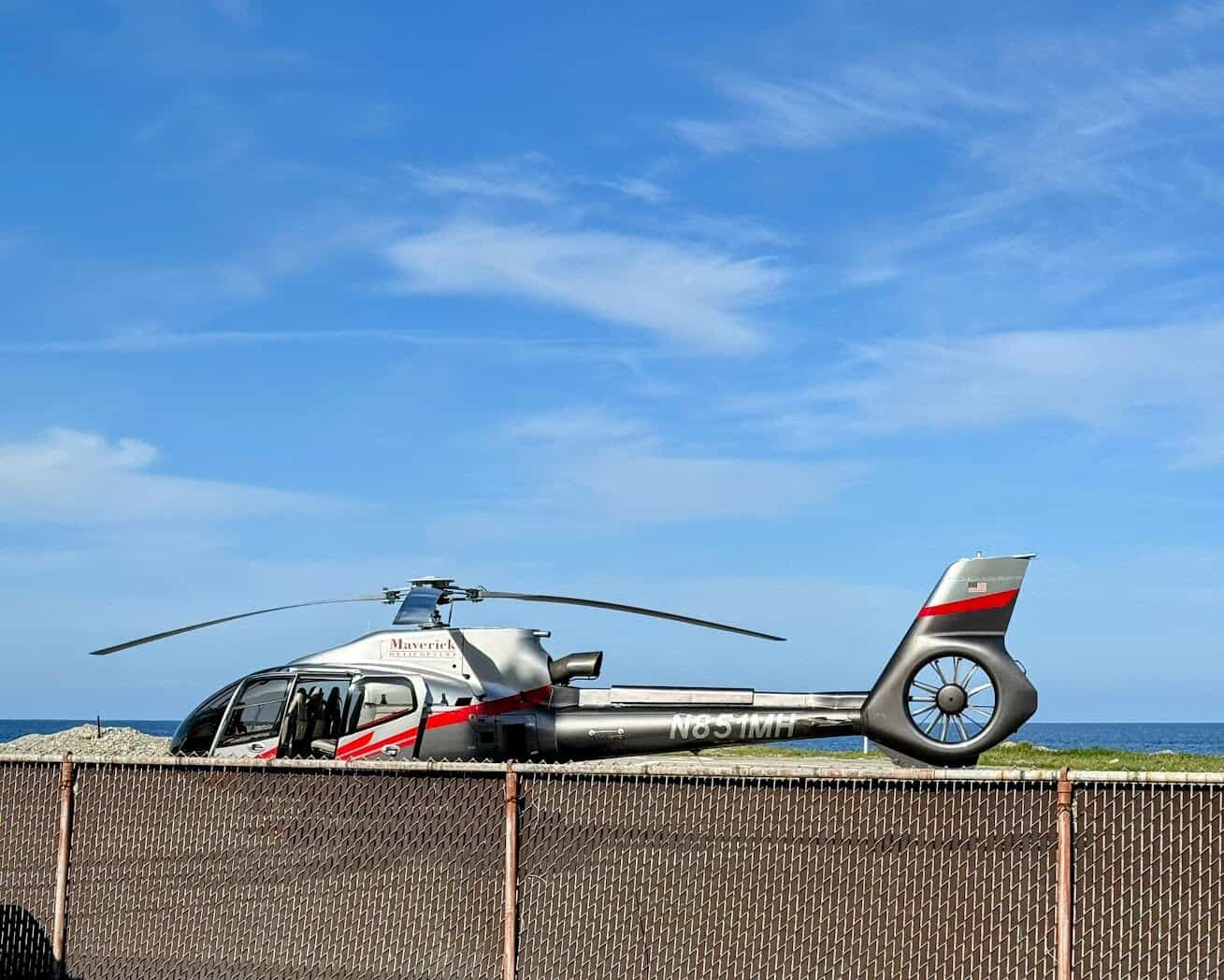 A Maverick helicopter with red and silver detailing is parked behind a chain-link fence, with the ocean visible in the background on Catalina Island.

